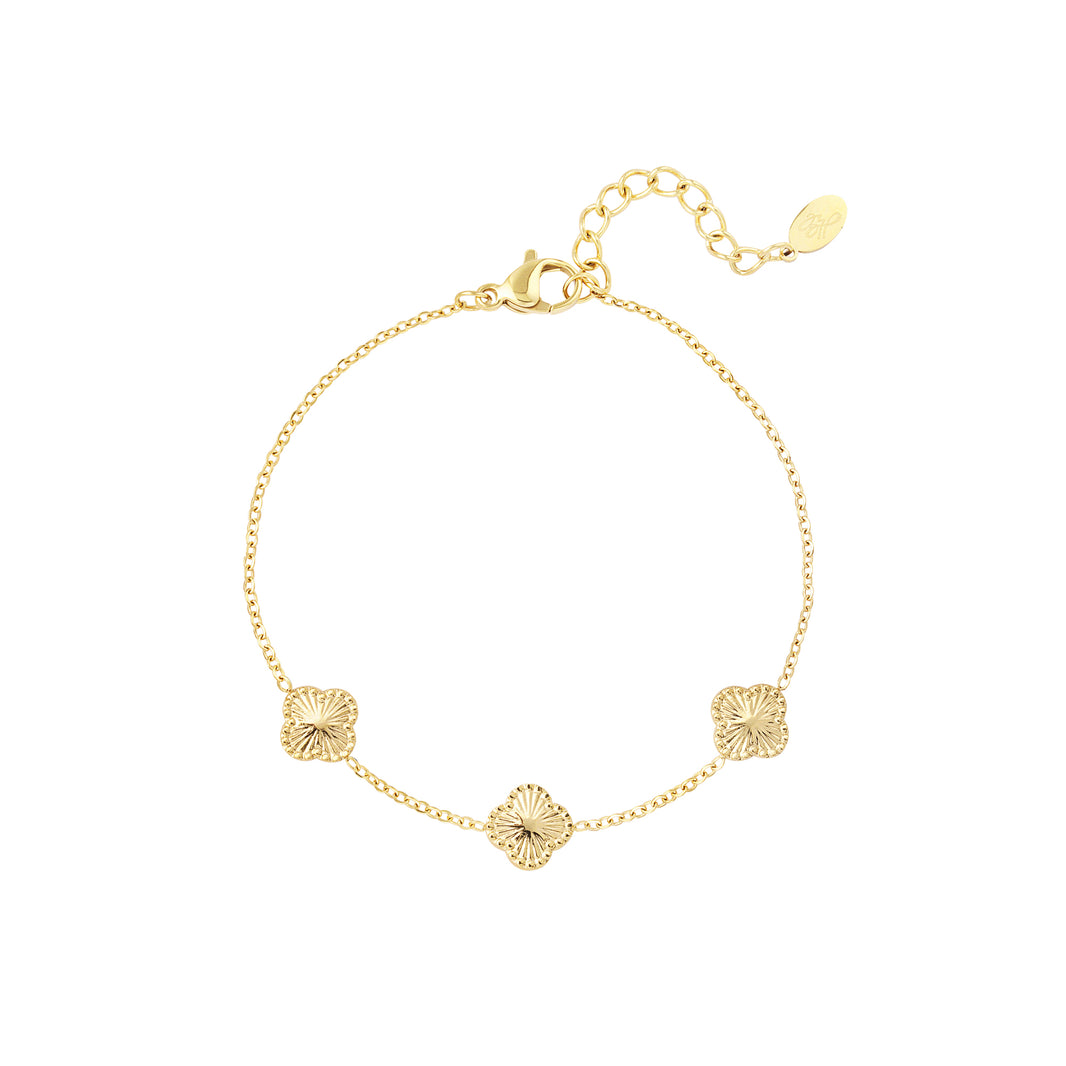 Das Bild zeigt ein filigranes Armband aus goldfarbenem Edelstahl, verziert mit drei stilisierten Kleeblatt-Elementen. Es liegt auf einem hellbeigen Hintergrund, der die warme Farbe des Schmucks elegant unterstreicht. Die zarten Glieder und die feine Struktur der Ornamente vermitteln Leichtigkeit und Qualität.