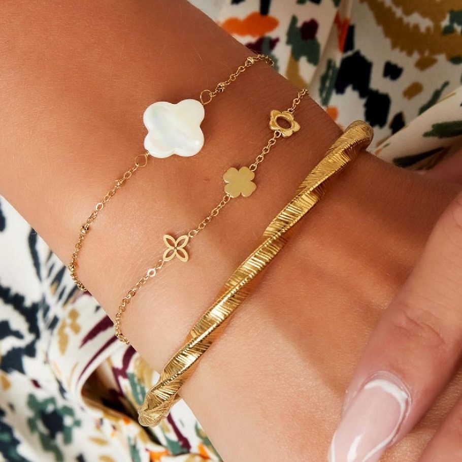 Nahaufnahme eines Handgelenks mit drei Armbändern, darunter das Mira Lou Armband aus goldfarbenem Edelstahl mit floralen Motiven