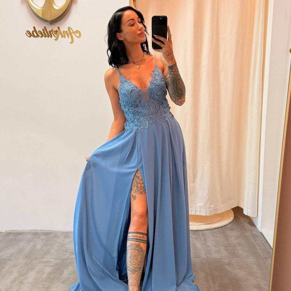 Model trägt das staubblaue Abendkleid „Lyanna“ mit floraler Spitze, Schlitz und zarten Trägern – ein femininer Look für besondere Anlässe.