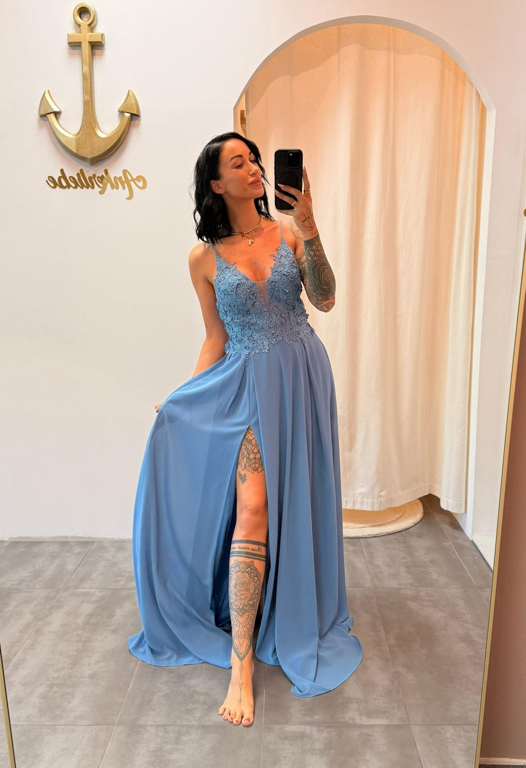 Model trägt das staubblaue Abendkleid „Lyanna“ mit floraler Spitze, Schlitz und zarten Trägern – ein femininer Look für besondere Anlässe.
