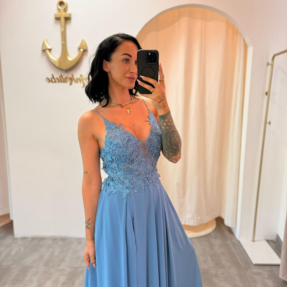 Model trägt das staubblaue Abendkleid „Lyanna“ mit floraler Spitze, Schlitz und zarten Trägern – ein femininer Look für besondere Anlässe.