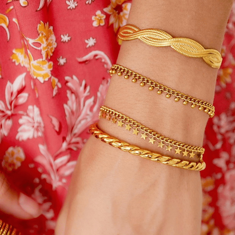 image Baila Armband am Handgelenk, kombiniert mit weiteren goldfarbenen Armbändern vor einem roten Blumenkleid