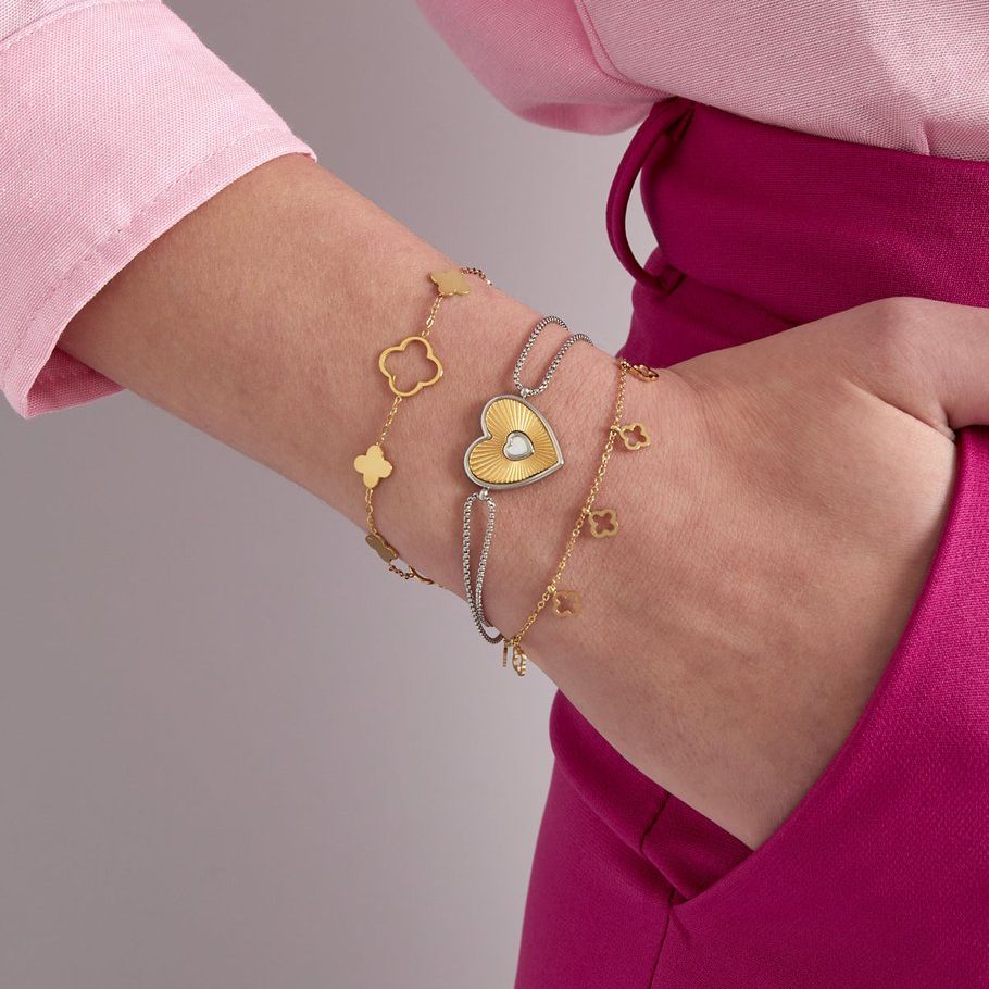 Goldene und silberne Armbänder mit Herz- und Kleeblattanhängern am Handgelenk, kombiniert mit pinker Hose